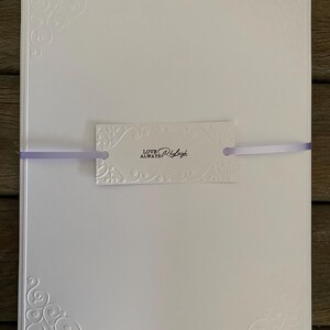 K&ouml;nnte beinhalten: Ein Stapel wei&szlig;er Schreibwaren mit gepr&auml;gten Eckdesigns und einem dekorativen Anh&auml;nger. Der Anh&auml;nger ist mit einem lavendelfarbenen Band gebunden und tr&auml;gt den Schriftzug "LOVE ALWAYS Ryleigh" in schwarzer Schrift.