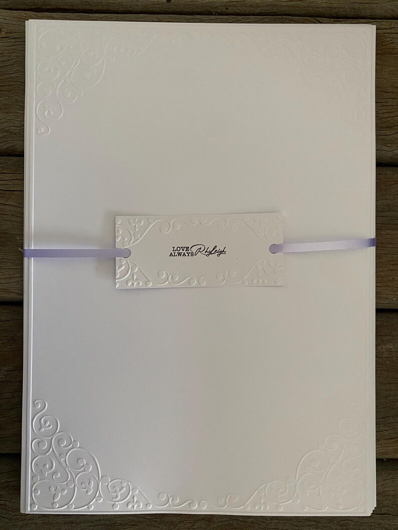 K&ouml;nnte beinhalten: Ein Stapel wei&szlig;er Schreibwaren mit gepr&auml;gten Eckdesigns und einem dekorativen Anh&auml;nger. Der Anh&auml;nger ist mit einem lavendelfarbenen Band gebunden und tr&auml;gt den Schriftzug "LOVE ALWAYS Ryleigh" in schwarzer Schrift.