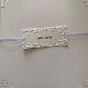 K&ouml;nnte beinhalten: Eine wei&szlig;e Karte mit gepr&auml;gten floralen Eckdesigns und einem dekorativen Anh&auml;nger. Der Anh&auml;nger mit dem Text "LOVE ALWAYS Rhyleigh" ist mit einem lavendelfarbenen Band befestigt. Die Karte hat eine strukturierte Oberfl&auml;che.