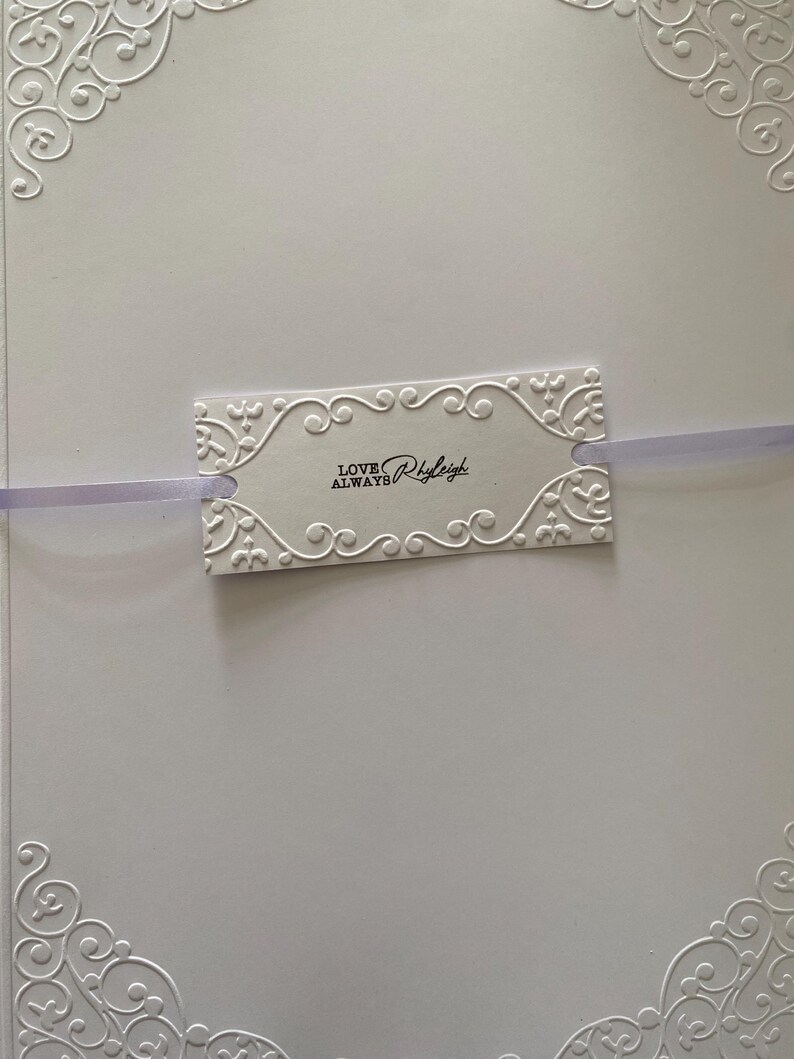 K&ouml;nnte beinhalten: Eine wei&szlig;e Karte mit gepr&auml;gten floralen Eckdesigns und einem dekorativen Anh&auml;nger. Der Anh&auml;nger mit dem Text "LOVE ALWAYS Rhyleigh" ist mit einem lavendelfarbenen Band befestigt. Die Karte hat eine strukturierte Oberfl&auml;che.