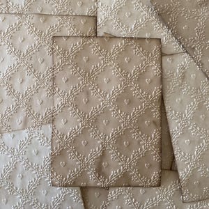 May include: Several beige paper sheets with an embossed floral and heart pattern. The design features a repeating lattice of leaves and small hearts, creating a textured surface. The paper has a vintage, aged appearance.