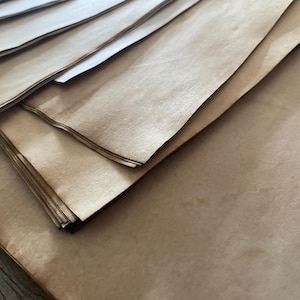 May include: A stack of rectangular sheets of paper with a light brown, tea-stained appearance. The paper has a smooth, matte texture with subtle variations in color, featuring darker edges and a slightly weathered, aged look across the surface.
