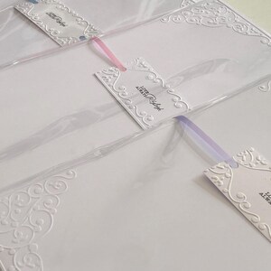 K&ouml;nnte beinhalten: Wei&szlig;e Schreibwaren mit gepr&auml;gten dekorativen Ecken und Anh&auml;ngern. Die Anh&auml;nger tragen den Text "Love Always Raleigh" und sind mit rosa und lila B&auml;ndern befestigt. Die Schreibwaren befinden sich in durchsichtigem Kunststoff.