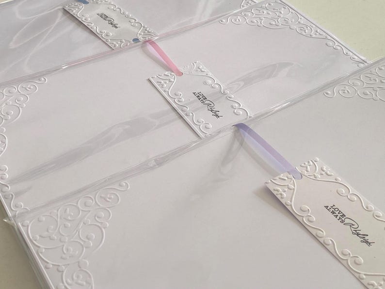 K&ouml;nnte beinhalten: Wei&szlig;e Schreibwaren mit gepr&auml;gten dekorativen Ecken und Anh&auml;ngern. Die Anh&auml;nger tragen den Text "Love Always Raleigh" und sind mit rosa und lila B&auml;ndern befestigt. Die Schreibwaren befinden sich in durchsichtigem Kunststoff.