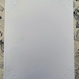 K&ouml;nnte beinhalten: Eine wei&szlig;e rechteckige Karte mit gepr&auml;gten floralen Eckendesigns. Die Karte hat eine saubere, minimalistische &Auml;sthetik mit komplizierten, wirbelnden Mustern in jeder Ecke. Der Hintergrund ist eine Vintage-Karte.