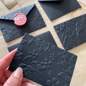 May include: Black stationery set featuring embossed floral designs. Includes envelopes, and cards. One envelope is sealed with a pink wax seal. The stationery is arranged on a neutral background.