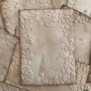 May include: A pile of aged paper sheets with embossed floral borders. The design features leaves and vines. The paper has a sepia tone with darker marks, giving a vintage appearance. The overall aesthetic is antique and reminiscent of old documents.