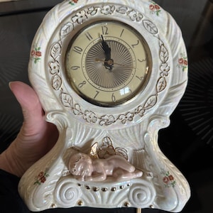 Op de afbeelding: Een witte keramische klok met een gouden rand en een cherub-ontwerp. De wijzerplaat van de klok heeft gouden Romeinse cijfers en een gouden uur- en minutenwijzer.