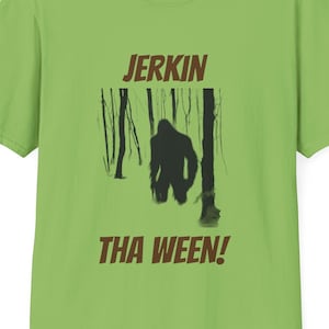 Puede incluir: Una camiseta verde con un gráfico marrón de una figura de Bigfoot en un bosque con el texto "Jerkin Tha Ween!" impreso en la parte delantera.