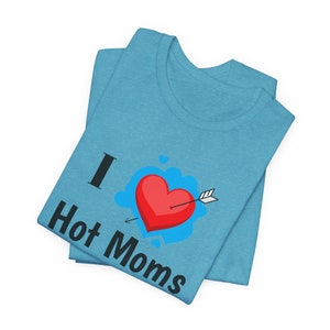 Puede incluir: Una camiseta azul claro doblada con el texto "I Hot Moms" en negro. Un corazón rojo atravesado por una flecha está en el centro, rodeado por un diseño de nube azul. La camiseta está hecha de un material suave.