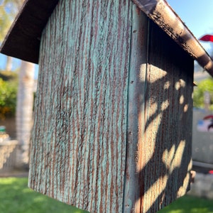 Op de afbeelding: Een klein houten vogelhuisje met een groene en bruine verweerde afwerking. Het vogelhuisje heeft een puntdak en hangt aan een haak.