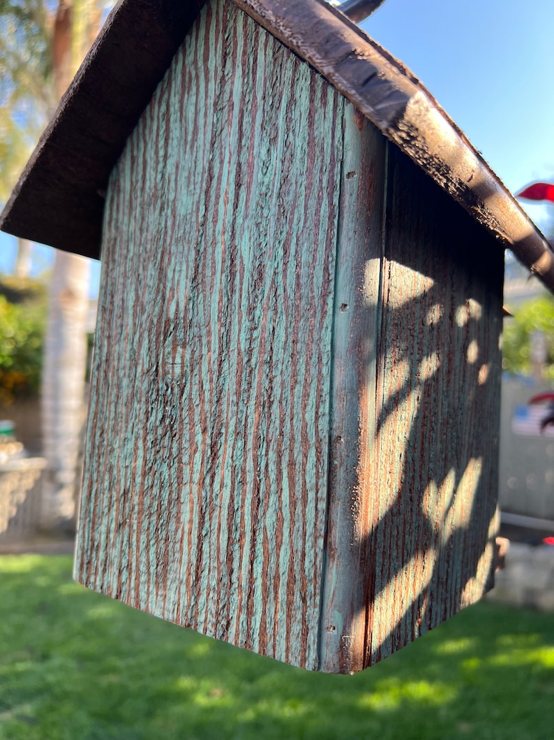 Op de afbeelding: Een klein houten vogelhuisje met een groene en bruine verweerde afwerking. Het vogelhuisje heeft een puntdak en hangt aan een haak.