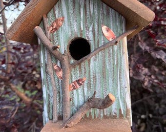 Op de afbeelding: Een houten vogelhuisje met een puntdak, voorzien van een lichtgroen geschilderd voorpaneel met verticale witte penseelstreken. De structuur is versierd met kleine natuurlijke takjes en diverse kleine, ovale, koperkleurige bladaccenten.