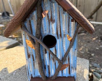 Op de afbeelding: Een rustiek vogelhuisje met een blauw-wit gestreept ontwerp, geaccentueerd met bruine houten afwerking en takken. Het vogelhuisje heeft een rond ingangsgat en is versierd met kleine, decoratieve bladeren. De basis is donkerbruin.