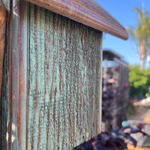 Op de afbeelding: Close-up van een houten vogelhuisje met een groene en bruine verweerde afwerking. Het vogelhuisje heeft een puntdak en een kleine opening waar vogels naar binnen kunnen.