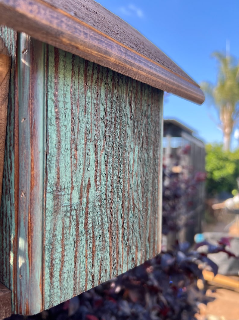 Op de afbeelding: Close-up van een houten vogelhuisje met een groene en bruine verweerde afwerking. Het vogelhuisje heeft een puntdak en een kleine opening waar vogels naar binnen kunnen.