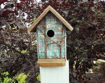 Op de afbeelding: Een houten vogelhuisje met een verweerde turquoise verfafwerking en een bruin zadeldak. De voorkant is voorzien van een rond invlieggat, een kleine rechthoekige zitstok en decoratieve natuurlijke takjes met kleine blaadjes op het oppervlak.