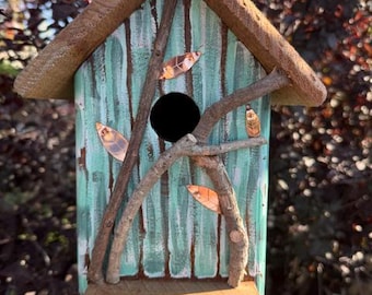 Op de afbeelding: Een houten vogelhuisje met een puntdak en een licht turquoise geschilderde voorkant met verticale, verweerde houtnerf. De structuur is versierd met natuurlijke bruine takjes en kleine koperkleurige bladaccenten rondom een centrale ronde opening.