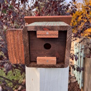 Op de afbeelding: Een houten vogelhuisje met een groen en blauw geschilderd exterieur. Het vogelhuisje heeft een kleine opening voor vogels om binnen te komen en een scharnierende deur aan de zijkant. Het vogelhuisje is gemonteerd op een witte houten paal.
