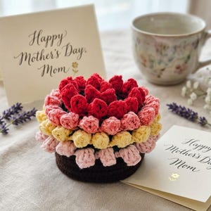 May include: A handmade crochet flower in shades of red, pink, and yellow sits on a table next to a teacup and a card that reads "Happy Mother's Day, Mom." The flower is made of multiple layers of crochet stitches.