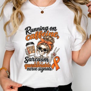May include: A white t-shirt featuring a graphic of a skeleton with a messy bun, orange polka-dot headband, and leopard-print sunglasses. The skeleton holds a coffee cup labeled "CAFFEINE: BECAUSE MS IS HARD" and makes a peace sign next to an orange ribbon.
