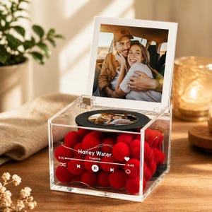 May include: A clear acrylic music box with a photo and red pom-poms inside. The box has a photo of a couple in a car and a small circular photo. The text "Honey Water" and a music player interface are visible.