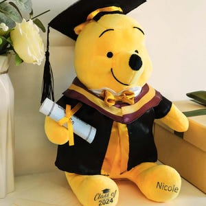 May include: A yellow plush Winnie the Pooh wearing a black graduation cap and gown with a maroon and gold trim. The bear holds a diploma and has "Class of 2024" and "Nicole" printed on its feet. A decorative graduation gift.