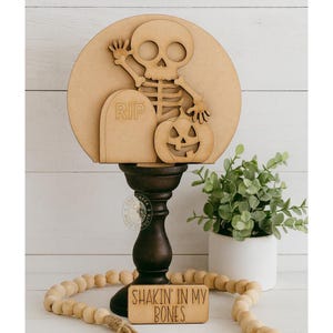 May include: A wooden Halloween-themed decoration featuring a skeleton, tombstone, and jack-o'-lantern on a round backdrop. The design is mounted on a dark brown candlestick base. A small wooden sign below reads "Shakin' in my Bones."