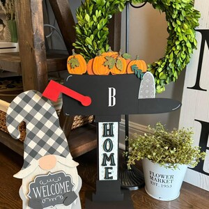 May include: A black and white checkered gnome holding a sign that says "Welcome".  A black mailbox with a red flag and pumpkins on top.  The mailbox is attached to a black stand with the word "HOME" in white letters.  A small white bucket with the text "FLOWER MARKET EST. 1953" is on the floor.