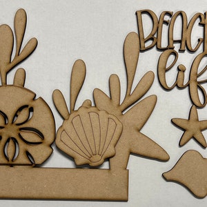 May include: Wooden cutout set with a beach theme. The set includes a seashell, starfish, sand dollar, seaweed, and a sign that reads "Beach Life".