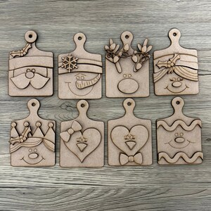 May include: Eight wooden ornaments shaped like cutting boards with various Christmas-themed faces, including Santa, a snowman, a reindeer, and a heart.