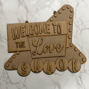 May include: Wooden sign with the text "Welcome to the Love Shack" in a retro style. The sign is brown and has a distressed look. It is perfect for adding a touch of whimsy to your home decor.