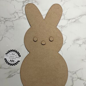 May include: A brown wooden cutout of a bunny rabbit with two black circles for eyes and a smaller black circle for a nose. The cutout is on a white marble background. The text "HANDCRAFTED PRODUCT ESTD 2017 DOG CREEK WOOD CUTOUTS CARPENTRY SERVICE" is on a black and white circle sticker.