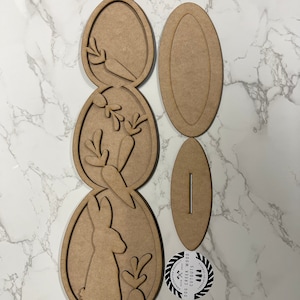 May include: A wooden craft kit for making an Easter bunny decoration. The kit includes three oval-shaped pieces with cutouts of a bunny, carrots, and leaves. There is also a separate piece for the base of the decoration. The kit is made by Dog Creek Wood Cutouts.