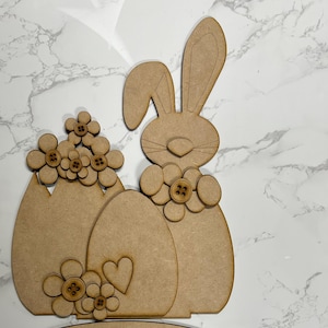 May include: Wooden craft kit featuring a bunny, two eggs, and flowers. The bunny has long ears and a cute face. The eggs are decorated with flowers. The kit is perfect for crafting and decorating for Easter.