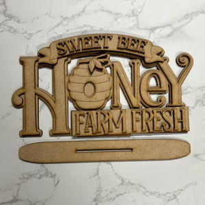 May include: A wooden sign with the words "SWEET BEE HONEY FARM FRESH" cut out in a decorative font. The word "HONEY" features a beehive graphic. The sign includes a base for standing.