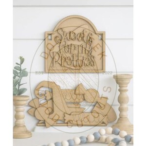 May include: Wooden sign with the text "Sweets for the Pretties" in a decorative font. The sign is light brown and has a carved design. The image also includes decorative wooden candle holders, a beaded garland, and a small plant.