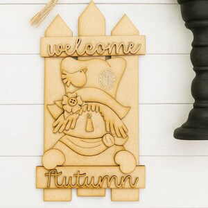 May include: Wooden welcome sign featuring a scarecrow design with the word "welcome" at the top and "autumn" at the bottom. The scarecrow has a hat, scarf, and flower. The sign is light brown and has a fence-like background.