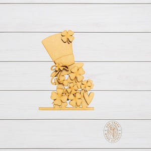 May include: A wooden cutout featuring a top hat and shamrocks, perfect for St. Patrick's Day. The hat is adorned with shamrocks and a bow. The cutout is made of light-colored wood and sits on a flat base.