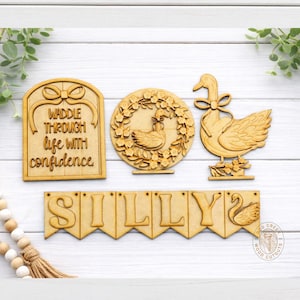 May include: Wooden cutouts featuring a swan theme. Includes a sign with the text "Waddle Through Life With Confidence", a circular wreath with a swan, a swan figure, and a banner spelling "SILLY".
