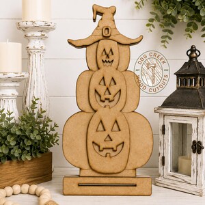 May include: A light brown wooden cutout featuring a stack of three jack-o'-lanterns topped with a witch's hat, all mounted on a rectangular base. The design includes cut-out facial features for each pumpkin and a circular logo with the text DOG CREEK WOOD CUTOUTS EST 2022.