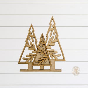 May include: Three wooden Christmas tree cutouts with leaf-like designs. The trees are arranged in a row, with the middle tree slightly smaller. The cutouts are made of light brown wood and are set against a white background.