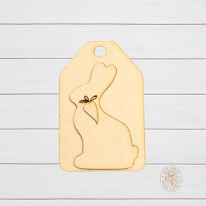 May include: A wooden tag with a laser-cut bunny design. The bunny holds a carrot and is tied with a bow. The tag has a hole at the top for hanging. The wood is unfinished and ready for painting or staining.
