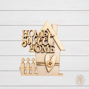 May include: A wooden cutout in the shape of a house with the words "HOME SWEET HOME" in a decorative font. The house features a heart with a button and a small picket fence. The cutout is made of unfinished wood.