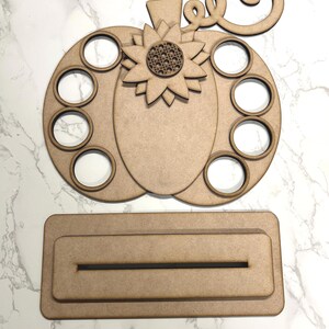 May include: Unfinished wooden pumpkin with eight circles cut out, a sunflower design, and a swirl on top. The pumpkin is on a rectangular base with a slot cut out.
