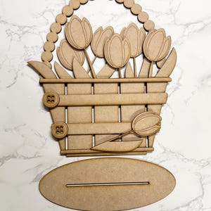 May include: Wooden cutout of a basket filled with tulips. The basket has a handle with a beaded edge and two buttons on the side. The basket is sitting on a separate wooden oval base.