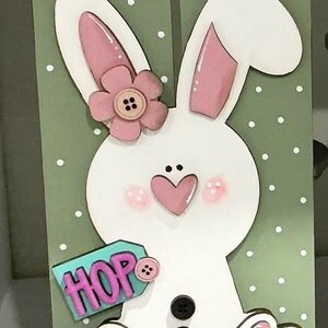 May include: A white bunny with pink ears and a pink flower on its head. The bunny is holding a teal tag with the word "HOP" in pink letters. The bunny is sitting on a green background with white polka dots.