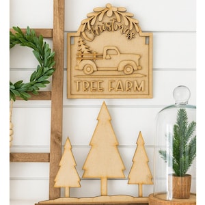 May include: Wooden Christmas sign with a truck carrying a Christmas tree, the words "Christmas" and "Tree Farm" are engraved. Three wooden Christmas trees are in front of the sign. A green wreath and a small tree in a glass dome are also visible.