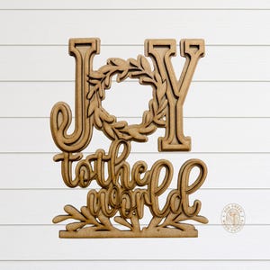 May include: Wooden sign with the words "JOY to the world" in a layered design. The word "JOY" is in large capital letters, with a wreath in the center. The phrase "to the world" is in cursive script. The sign is made of unfinished wood.