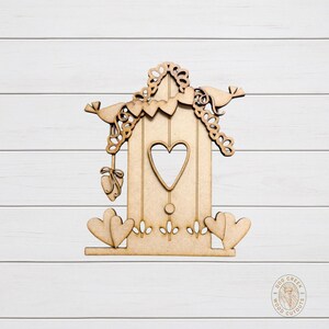 May include: A wooden birdhouse cutout with a heart-shaped opening and decorative elements. The birdhouse features a heart-shaped cutout, two birds, and a hanging heart-shaped ornament. Two heart shapes are at the base. The cutout is made of light brown wood.